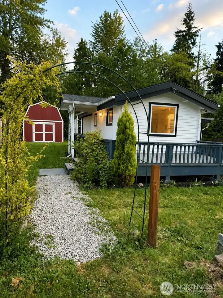 Welcome to your own private retreat! This inviting backyard features a picturesque gravel pathway framed by a graceful green garden arch draped in lush greenery. It is Peaceful backyard with garden arch, gravel path, large deck, and iconic red barn — the perfect blend of charm and outdoor living space.