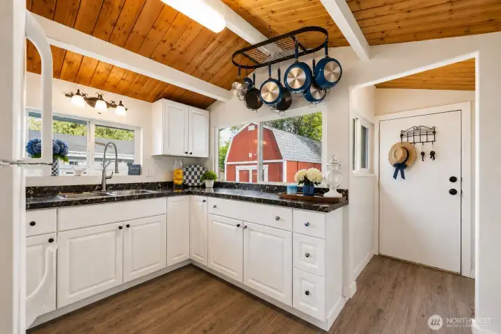 Bathed in abundant natural light, this beautifully updated kitchen is a true highlight of the home. Featuring crisp white cabinetry, sleek dark granite countertops, and warm hardwood-style flooring, the space offers both style and functionality. The generous corner sink area with a modern gooseneck faucet sits beneath large windows that frame stunning views of the lush backyard and iconic red barn. Vaulted tongue-and-groove wood ceilings with white beams add dramatic character and height, while the stainless steel appliances and thoughtful layout create an efficient, bright workspace perfect for everyday cooking and entertaining. This inviting kitchen seamlessly blends modern updates with the home’s classic Craftsman charm.