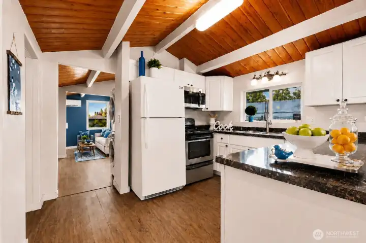 This bright and inviting kitchen is a true centerpiece of the home, featuring crisp white cabinetry, sleek dark granite countertops, and warm wood-look flooring beneath soaring vaulted tongue-and-groove cedar ceilings with exposed white beams. The well-equipped space includes a full-size refrigerator, stainless steel range and oven, generous counter workspace, and a convenient peninsula perfect for casual meals or entertaining. Abundant natural light pours in through large windows overlooking the backyard, while modern lighting and thoughtful finishes create a functional, cheerful atmosphere ideal for everyday cooking and family gatherings. Perfectly blending classic charm with practical updates, this kitchen offers everything you need to create lasting memories.