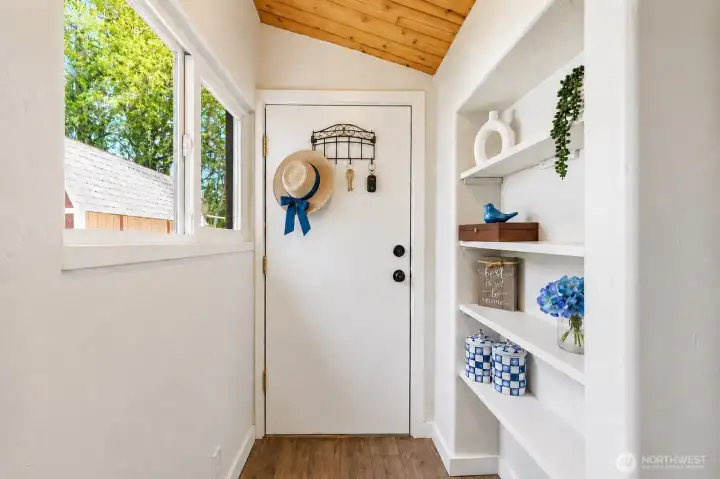 This bright and functional rear entryway provides direct access to the backyard through a convenient white door. Featuring the home’s signature vaulted tongue-and-groove wood ceiling, the space is enhanced with custom built-in shelving ideal for storage, coats, shoes, and everyday items. A large window fills the area with natural light, while thoughtful details like the key rack and organized shelving make this the perfect drop zone when coming in from the backyard, deck, or garden. Practical, stylish, and welcoming, this rear entrance adds everyday convenience and charm.