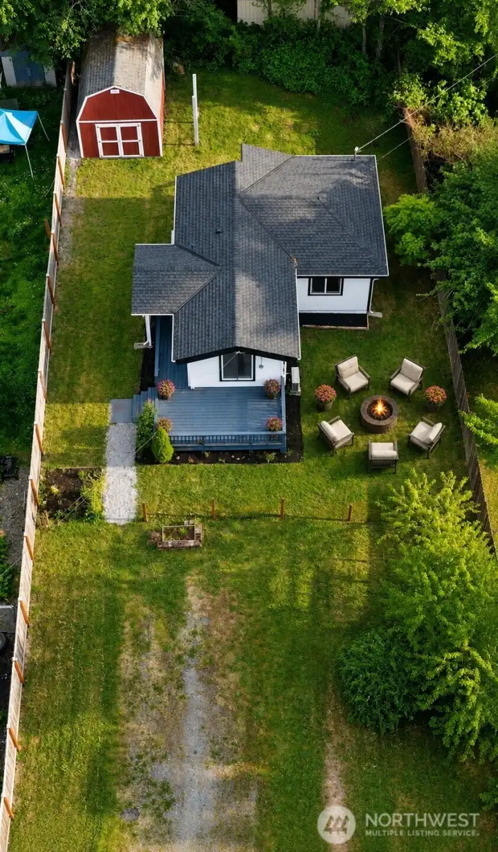 This aerial view beautifully captures the expansive private lot of this charming home, showcasing a spacious, level backyard with lush green lawn, mature trees for natural privacy, and excellent outdoor living potential. The generous gravel driveway provides ample on-site parking with plenty of room for an RV and multiple vehicles. A large red storage shed with double-door access and lofted storage areas adds significant utility, while the home’s welcoming blue deck extends seamlessly into the yard for effortless indoor-outdoor enjoyment. Tucked at the end of a quiet dead-end street, the property combines privacy, functionality, and plenty of room to garden, entertain, or simply relax in a serene, tree-lined setting.