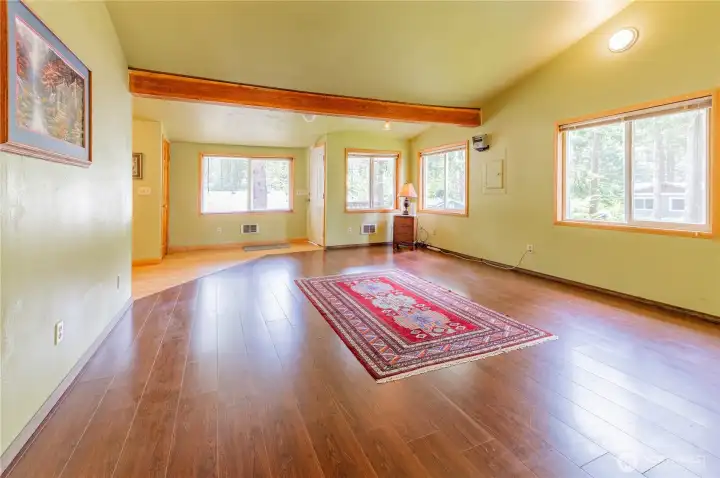 Spacious main living area with vaulted ceilings, exposed beam, hardwood-look flooring, and large windows bringing the forest indoors.