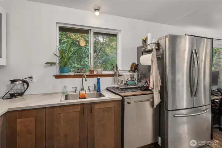 Unit 1 Large picture window above the sink brings natural light and views of mature greenery, paired with stainless-steel dishwasher and solid surface countertops.