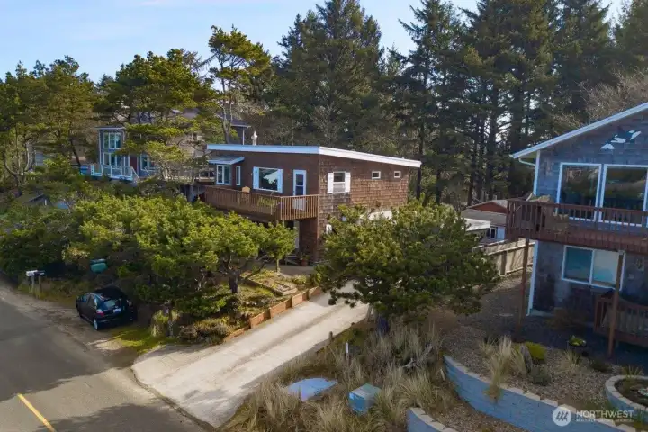 This elevated view showcases the home’s wooded coastal setting, highlighting its upper-level deck and welcoming presence among mature evergreens.