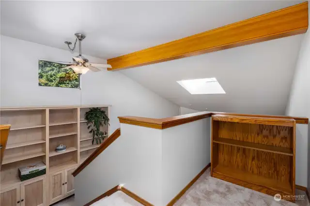 Wide carpeted stairway leads to upper level with ceiling fan, built in bookshelves at mid landing, and windows overlooking living room