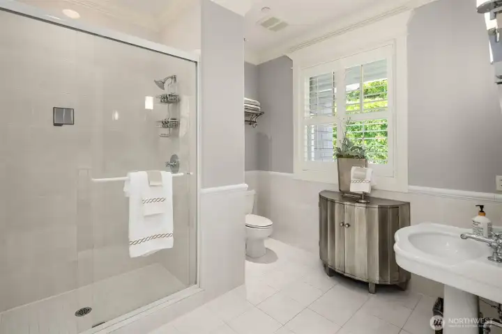 This beautifully finished main floor guest bathroom serves as a private ensuite for the main-level guest quarters. The bright and functional 3/4 bath features a classic pedestal sink, an elegant contemporary vanity console for storage, a seamless glass-enclosed step-in shower, and a large window equipped with plantation shutters that flood the space with soft, natural light. This beautifully finished main floor guest bathroom serves as a private ensuite for the main-level guest quarters. The bright and functional 3/4 bath features a classic pedestal sink, an elegant contemporary vanity console for storage, a seamless glass-enclosed step-in shower, and a large window equipped with plantation shutters that flood the space with soft, natural light.