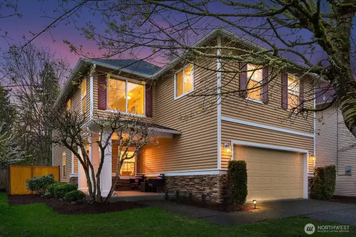 This alternate twilight view of the home's exterior showcases the warm glow of interior lights illuminating the classic two-story profile against a beautiful dusky sky, highlighting the home's inviting presence and well-maintained landscaping. The charming stone accents, two-car garage, and lush mature plantings create a picture-perfect curb appeal that is equally stunning day or night.