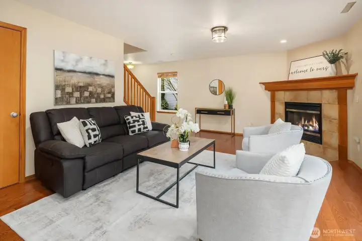This cozy view of the living room showcases the charming fireplace with its warm stone surround alongside the rich hardwood floors and designer paint that give the space its welcoming character. The open sightline to the staircase and the natural light filtering throughout create a bright, connected, and comfortable atmosphere that is perfect for everyday family living.