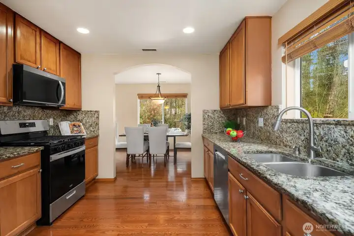 This alternate view of the kitchen highlights the generous layout with ample counter space, ceiling-high cabinetry, brand new stainless steel appliances including an over-the-range microwave, and beautiful granite countertops that tie the space together with a polished, elevated aesthetic. The hardwood floors and open flow toward the bright dining area beyond make this a wonderfully functional and inviting space for everyday cooking and entertaining.