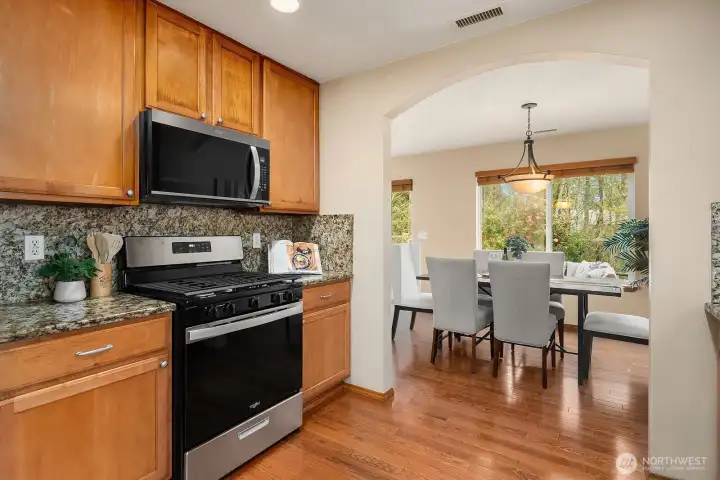 A closer look at the kitchen showcases the brand new stainless steel oven range and over-the-range microwave, beautiful granite countertops with elegant tile backsplash, and the warm ceiling-high cabinetry that provides exceptional storage throughout. The seamless sightline through the arched opening to the sun-filled dining room beyond perfectly illustrates the thoughtful open-concept design of this well-appointed home.