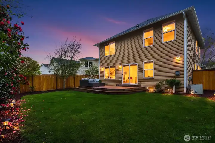 A stunning twilight view of the home's back exterior, with warm light glowing from within and the deck beautifully illuminated — showcasing the perfect blend of indoor and outdoor living at golden hour.