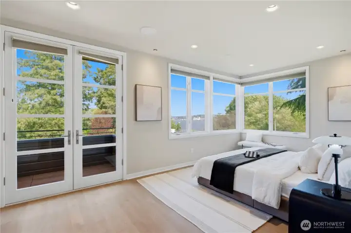 Spacious primary with a fireplace set into custom walnut built-ins, French doors to a private balcony, and large picture windows overlooking the blue and green.