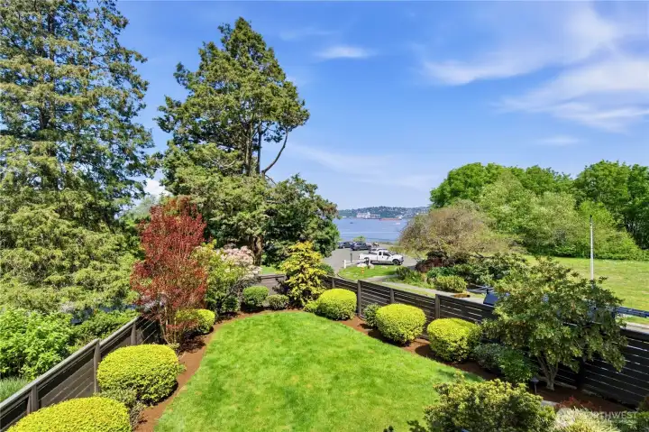 A professionally landscaped rear garden with manicured lawn, Japanese maple, and blooming rhododendrons — with the Sound peeking through at the back.