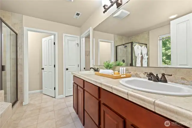 Luxurious primary bathroom with double vanity and walk-in shower in Puyallup, WA    Enjoy spa-like comfort in this spacious primary ensuite bathroom, featuring a dual-sink tile countertop vanity, private water closet, a large walk-in shower, and natural light from a window overlooking the greenbelt. This tranquil retreat is located upstairs in the highly sought-after LaGrande Station community of Puyallup, WA, just minutes from the Washington State Fairgrounds.