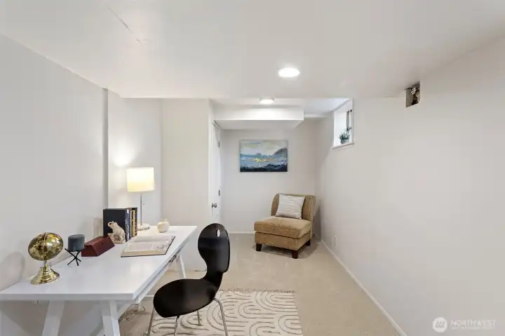 A long lower-level flex room with two west-facing windows for natural light, alternate laundry hookups, and versatility for use as a media room, office, studio, or workshop, with access to storage and the mechanical area beyond.