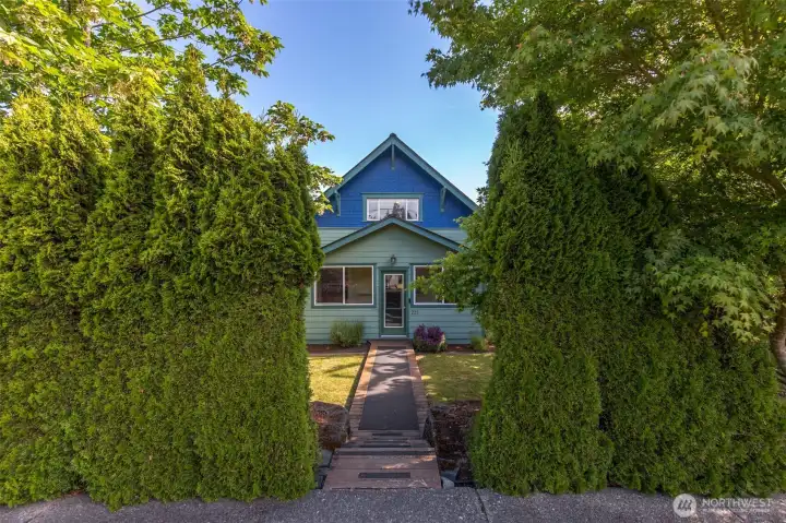 A symmetrical front view of the home framed by tall, manicured privacy hedges with a walkway leading up to the charming blue exterior.