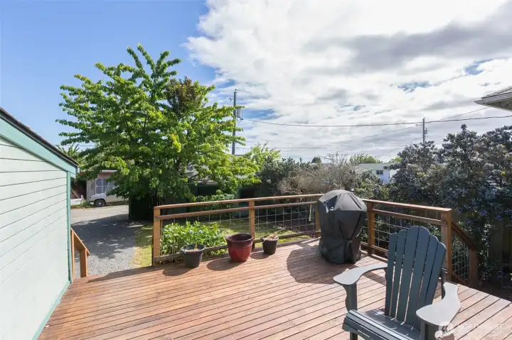 A view from the refinished wooden deck overlooks the gravel parking area and surrounding lush green trees.