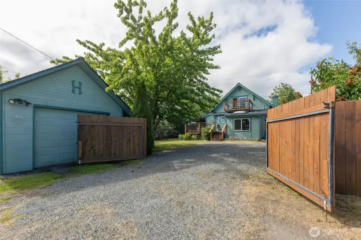 An open wooden gate reveals the gravel driveway and extra off-alley parking leading toward the detached garage and main house.