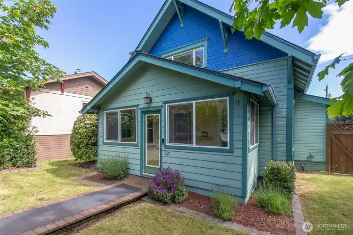 A wooden walkway leads past landscaped garden beds to a cozy side entrance of the charming blue home.
