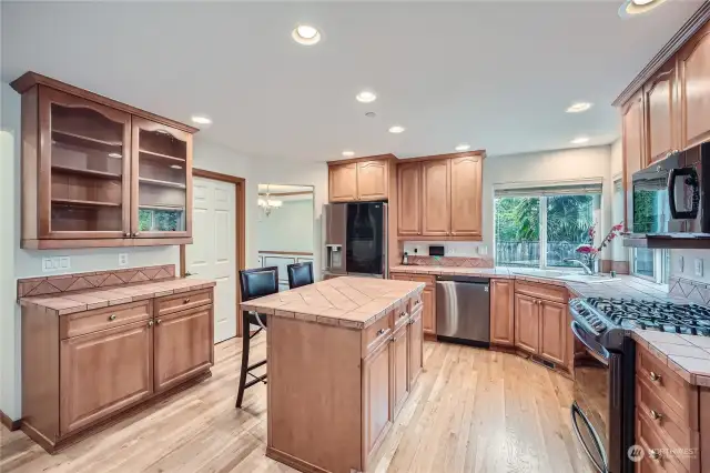 This kitchen features warm wood cabinetry that offers ample storage, with an island at its center for additional workspace. The countertops have a tiled design that extends into a backsplash, providing a rustic, earthy look. Recessed lighting illuminates the space, giving it a bright and inviting feel. Stainless steel appliances, including a gas range, dishwasher, and modern refrigerator, add a sleek contrast to the cabinetry. A large window over the sink brings in natural light and offers a view of the backyard. The hardwood floors enhance the warmth and cohesion of the room, while the open layout gives it a spacious flow.