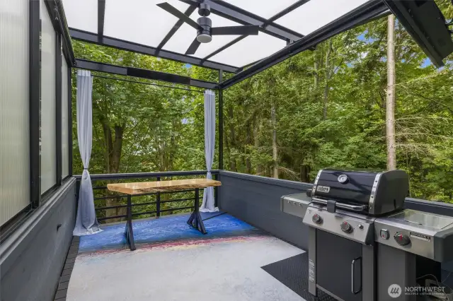 Outdoor living space overlooks trees, features dual heaters, ceiling fan and pull down shades for a comfortable space year-round