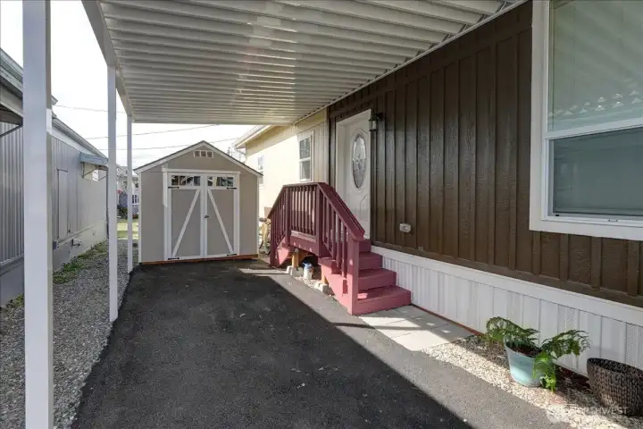 This home features a practical and covered outdoor area, offering protection from the elements year-round. The space provides easy access to a dedicated storage shed, perfect for keeping tools, bikes, and other outdoor gear organized and out of sight. A set of steps with a handrail leads to the home's entrance, providing both accessibility and a welcoming feel. This functional design makes outdoor living simple and tidy.