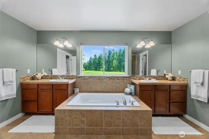 A soaking tub is nestled between the separate vanities with an incredible view of the golf course and evergreen trees in this primary bathroom... a PNW treat!
