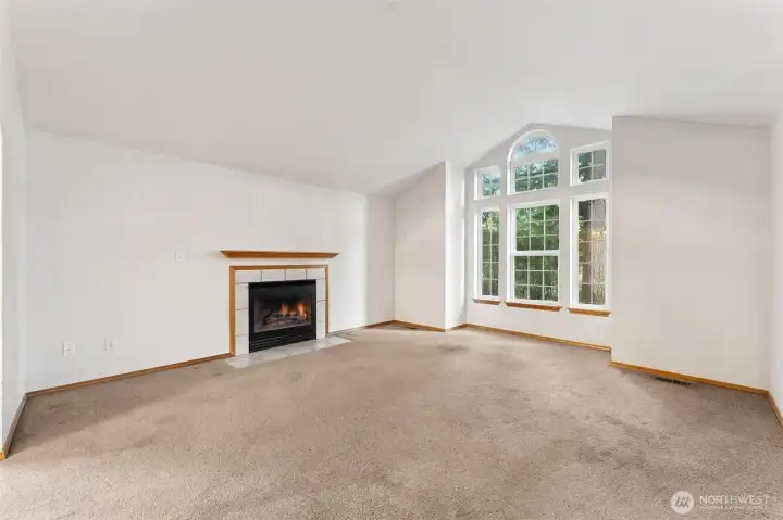 The living room opens up in a big way with vaulted ceilings and a wall of windows that immediately grabs your attention. Six windows stack together from floor to ceiling, with portrait panes below and an arched accent above, framed by decorative grilles that give them a classic look. The light pours in all day and makes the whole space feel bright and welcoming. A gas fireplace with a simple tile surround and clean mantle anchors the room, giving it a cozy focal point in a space that easily handles full size furniture and gatherings.