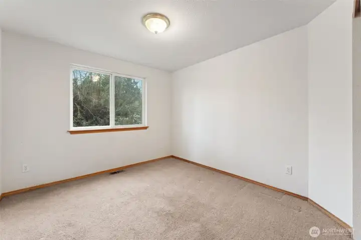 A little larger than the previous room, this bedroom leans simple and practical with white walls, neutral carpet, and those warm wood trim details around the baseboards and window sills. The natural wood gives the space a softer feel than stark white trim and pairs nicely with just about any furniture. Easy to picture as a bedroom, guest space, or a very pleasant work from home spot.