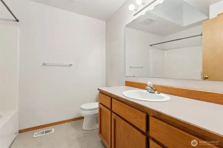 The main bathroom keeps things bright and functional with white walls, a long oak shaker vanity, and a wide landscape mirror that makes the space feel larger right away. Vinyl flooring handles daily traffic easily, and the full tub and shower combo keeps routines simple. The skylight is the quiet star here, pulling in natural light so mornings are just a bit brighter.