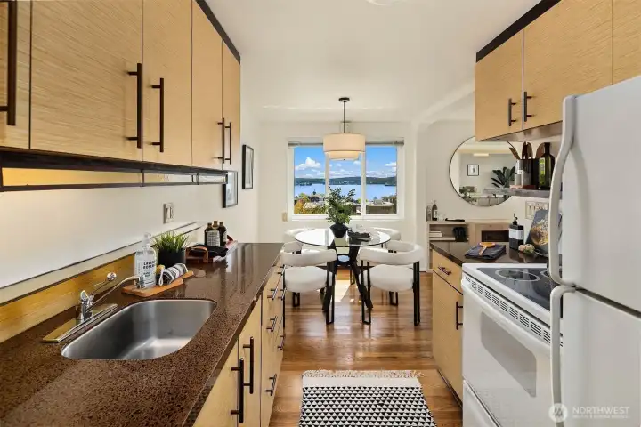 The kitchen looking toward the dining area — and wow, those views from the 6th floor! Elliott Bay and the Puget Sound shimmer straight ahead. Updated cabinets with black hardware, dark granite-look countertops, dishwasher, and hardwood floors make this a kitchen that works as hard as the scenery.