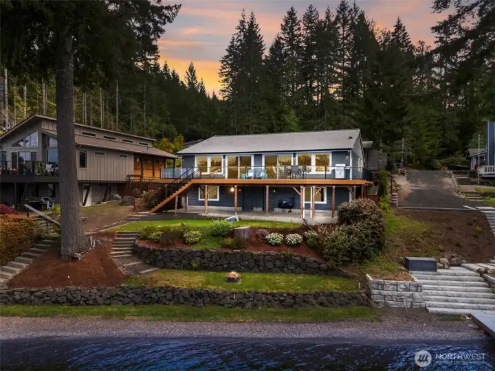 From out over the water, this twilight aerial captures the full layered beauty of the property, 60 feet of pebble beach shoreline, terraced lawn areas, landscaped beds and the dramatic deck and patio spaces rising naturally back toward the home, and the design here is smart as well as beautiful. Wide, easy-access steps connect each level comfortably, making the waterfront feel inviting and usable instead of steep or complicated. The entire setting glows at dusk, with lights reflecting softly against the home while the lake settles into that glassy evening calm people dream about when they picture waterfront living. It feels peaceful, elevated and just a little magical.