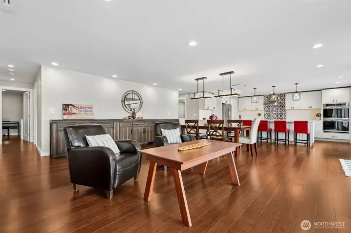The entire back wall of the home opens to Mason Lake through dramatic near floor-to-ceiling windows, pulling the water, sky and sunset views straight into the living space like living artwork. Those wide plank coffee-toned hardwood floors stretch through the entire open-concept layout, connecting the living room, second sitting area, dining space and kitchen into one long, effortless flow built for entertaining, weekend guests or simply living really well every day.