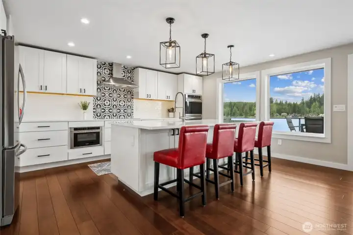 Now this kitchen has main character energy. That oversized island with seating for at least four sits right beside sweeping Mason Lake views, which means even grabbing coffee or answering emails suddenly feels suspiciously scenic while three contemporary pendant lights bring the perfect amount of visual punch overhead. Crisp white shaker cabinetry, black hardware, creamy subway tile and that dramatic counter-to-ceiling accent wall behind the stainless hood all layer together beautifully, and the inset microwave tucked into the lower cabinetry? Thank you. No giant microwave awkwardly hanging over the stove ruining the vibe.