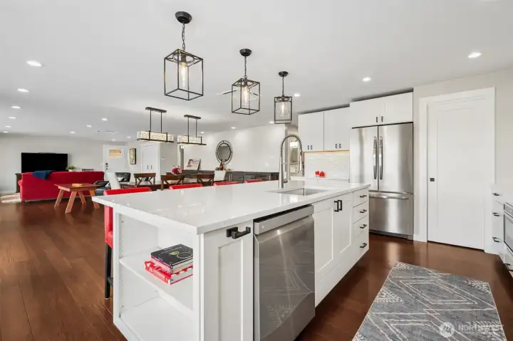 The built-in shelving detail on the end gives it personality and practicality at the same time, adding a custom furniture feel instead of just another oversized slab in the middle of the room. The large undermount single-basin sink and commercial-style pull-down faucet lean slightly chef-y in the best possible way, ready for entertaining, cleanup or someone dramatically rinsing wine glasses while staring out at the lake. White quartz counters keep the palette bright and elevated, while those rich coffee-toned wide plank hardwood floors continue seamlessly through the kitchen, dining and living areas, tying the entire waterfront space together beautifully.