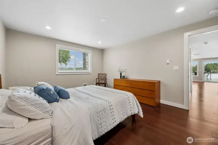 This primary suite has a really lovely connection to the rest of the home while still feeling tucked into its own comfortable retreat. From the doorway you can see straight through toward the living and dining spaces and all the way out to the sliding glass doors beyond, giving the room an airy, connected feel instead of feeling boxed away down some isolated hallway. At approximately 12x15, it easily handles a king-sized bed with room left over for a seating area, reading corner or remote workspace setup if life occasionally follows you to the lake. The additional window keeps natural light flowing in, while the soft neutral palette and warm hardwoods continue the calm, cohesive feeling carried throughout the home.