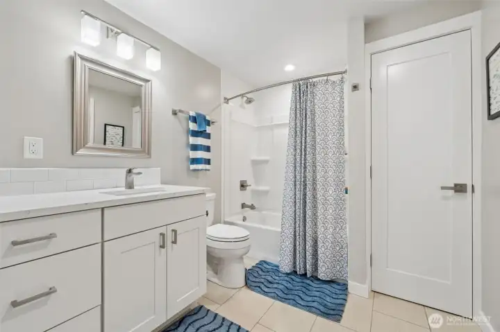The long white shaker vanity gives this space a clean, timeless look while offering generous counter space that people always appreciate once toothbrushes, skincare and random products start multiplying. The undermount sink, framed mirror and warm cream-toned tile floors keep everything feeling cohesive and elevated without tipping into trendy-for-the-sake-of-trendy territory. There is also a linen closet tucked in here for extra storage, because lake houses somehow require double the towels at all times, and the additional doorway leading directly to the laundry room adds a really smart layer of convenience to the floor plan.