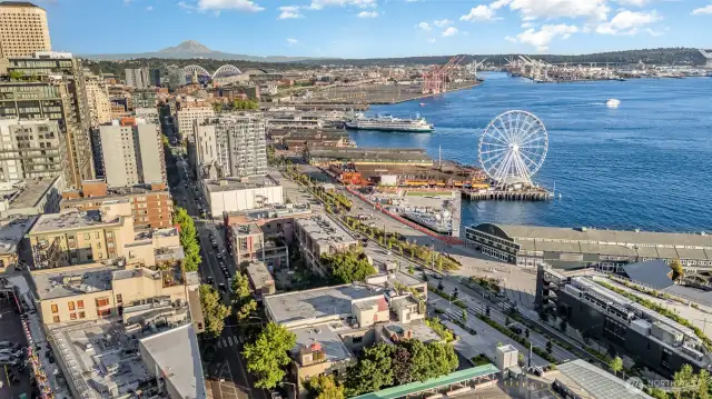 Marvel at this stunning aerial view from your Seattle waterfront condo, featuring the majestic Mount Rainier, the iconic Great Wheel, and a bustling ferry terminal against a vibrant blue Puget Sound. The photo captures the lively downtown skyline, lush greenery, and the bustling port, all within steps of your historic 1910 Fix Madore building. Enjoy unparalleled access to scenic water vistas and urban excitement, with the Ferris wheel adding a playful charm to your everyday panorama!