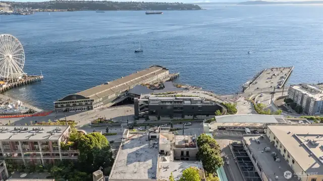 Capture the essence of Seattle waterfront living with this striking aerial view, featuring the shimmering Puget Sound, the iconic Great Wheel, and distant cargo ships dotting the horizon. The photo showcases the building's prime location near bustling piers and the Seattle Aquarium, with lush greenery and scenic water vistas enhancing the urban landscape. Enjoy unparalleled proximity to this vibrant waterfront hub, where every glance offers a picturesque blend of nature and city life!