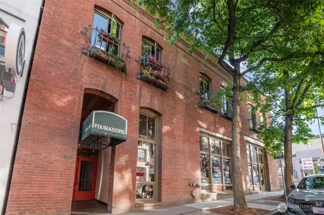 Step into historic charm with this stunning view of the 1910 Fix Madore building, its rich red brick facade adorned with vibrant flower boxes and arched windows. The photo captures the inviting entrance at 1507 Western Ave, framed by lush green trees, exuding a warm, welcoming vibe. Nestled in the heart of Seattle’s waterfront district, this pet-friendly condo promises a blend of classic architecture and urban convenience, just steps from Pike Place Market and the new Waterfront Park!