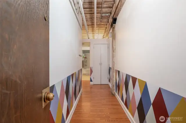 Step into a vibrant urban retreat with this inviting hallway, featuring warm hardwood floors and a striking geometric wall design in bold hues. The photo showcases exposed wooden beams and ceiling, adding historic charm, while a glimpse of the modern bathroom hints at updated comforts. This 815 sq ft condo blends classic architecture with contemporary flair, offering a welcoming entry to your stylish Seattle waterfront home!