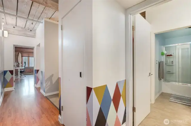 Welcome to a stylish retreat with this bright hallway, featuring warm hardwood floors and bold geometric wall accents in vibrant colors. The photo reveals exposed wooden beams and ceiling, blending historic charm with modern updates, while an open door offers a peek at the sleek bathroom with a glass shower. This 815 sq ft condo combines artistic flair and functional design, creating an inviting entry to your Seattle waterfront haven!