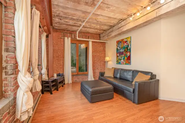 Experience versatile living in this spacious 815 sq ft condo, featuring a stylish living area with warm hardwood floors and exposed brick walls. The photo highlights a modern workspace  complemented by large windows with natural light. Exposed wooden beams and steel accents add historic charm, making this Seattle waterfront retreat perfect for work and relaxation!