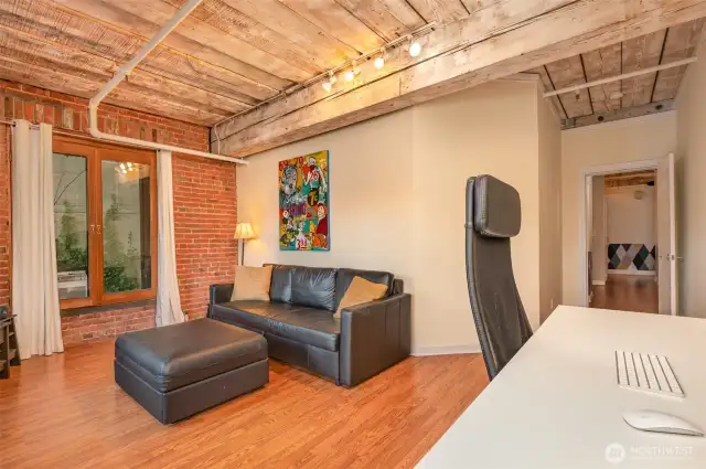 Experience versatile living in this spacious 815 sq ft condo, featuring a stylish living area with warm hardwood floors and exposed brick walls. The photo highlights a modern workspace  complemented by large windows with natural light. Exposed wooden beams and steel accents add historic charm, making this Seattle waterfront retreat perfect for work and relaxation!