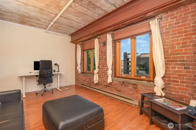 Experience versatile living in this spacious 815 sq ft condo, featuring a stylish living area with warm hardwood floors and exposed brick walls. The photo highlights a modern workspace  complemented by large windows with natural light. Exposed wooden beams and steel accents add historic charm, making this Seattle waterfront retreat perfect for work and relaxation!