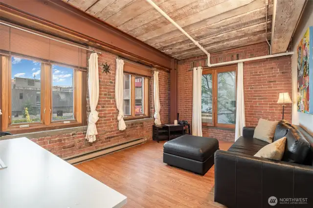 Experience versatile living in this spacious 815 sq ft condo, featuring a stylish living area with warm hardwood floors and exposed brick walls. The photo highlights a modern workspace  complemented by large windows with natural light. Exposed wooden beams and steel accents add historic charm, making this Seattle waterfront retreat perfect for work and relaxation!