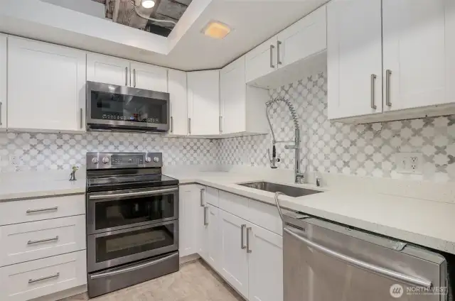 Whip up culinary delights in this modern kitchen, featuring sleek white cabinetry, a stylish mosaic tile backsplash, and a Samsung refrigerator. The photo highlights a spacious quartz countertop, stainless steel appliances including a double oven, and exposed wooden beams for a touch of historic charm.