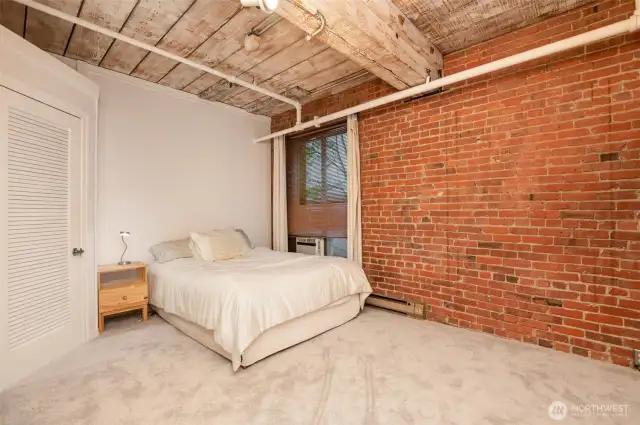 Unwind in this serene 1-bedroom retreat, featuring a plush carpeted floor and a cozy bed with soft linens. The photo highlights exposed brick walls and wooden beams, adding historic charm, while large windows with A/C offer natural light and comfort.
