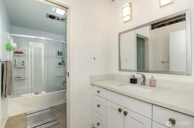 Refresh in this modern 1-bathroom, featuring a sleek quartz countertop and white cabinetry with ample storage. The photo showcases a spacious vanity with a large mirror, complemented by a glass-enclosed shower and tub combo, with stylish lighting and a clean design.
