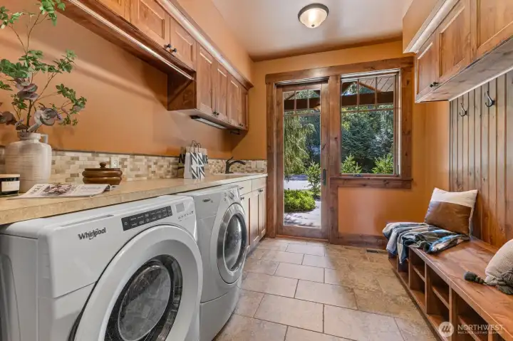 Light-filled laundry/mud room with private entrance, extensive knotty-pine cabinetry, travertine floors, and deep utility sink — ideal for pet care, gardening cleanup, or household projects. Includes laundry chute, shoe cubbies, and abundant storage for organized living.